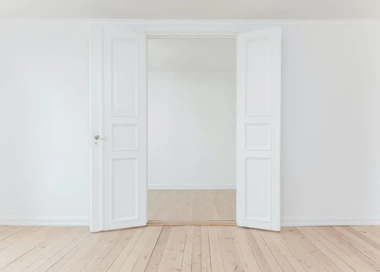 Otevřené bílé dveře vedou do prázdné místnosti s dřevěnou podlahou.