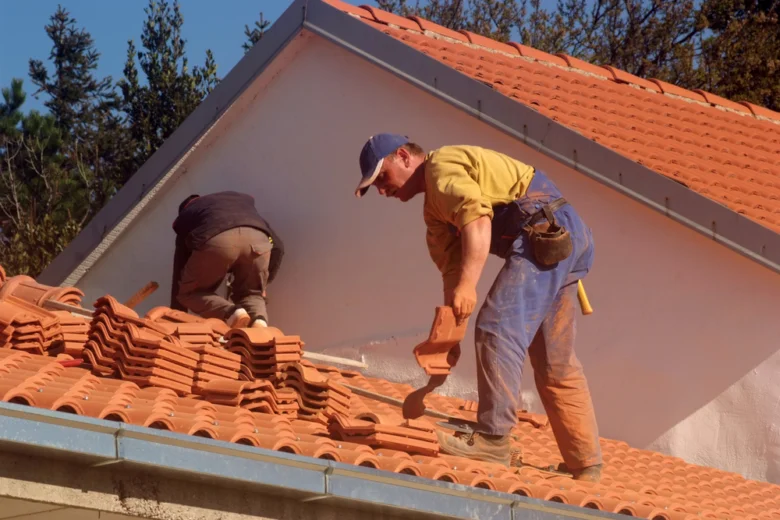 Dva dělníci montují červené střešní tašky na skloněnou střechu.