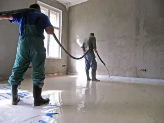 Dva dělníci nalévají betonový potěr do nezařízené místnosti.