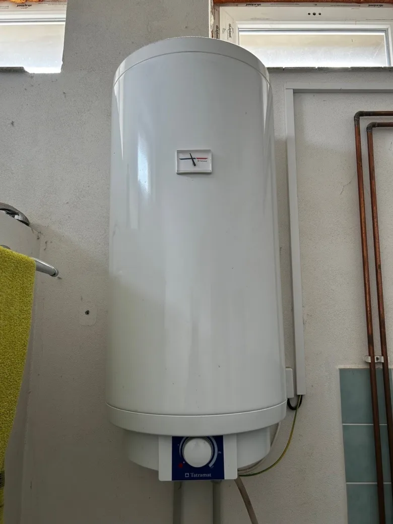 Bílý elektrický bojler značky Tatramat na zdi s měřidlem teploty.
