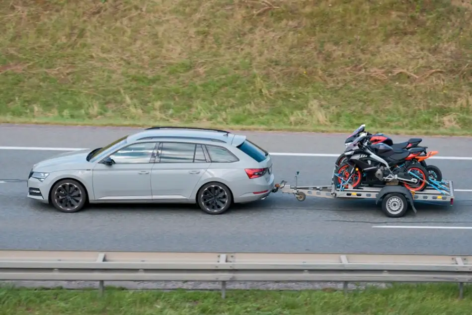Stříbrný vůz táhne přívěsný vozík s motocykly na dálnici.