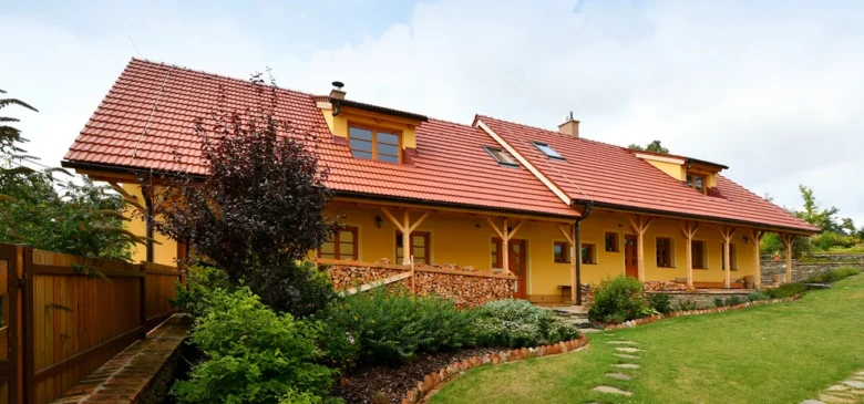 Žlutý dům s červenohnědou keramickou střechou v zahradním prostředí.