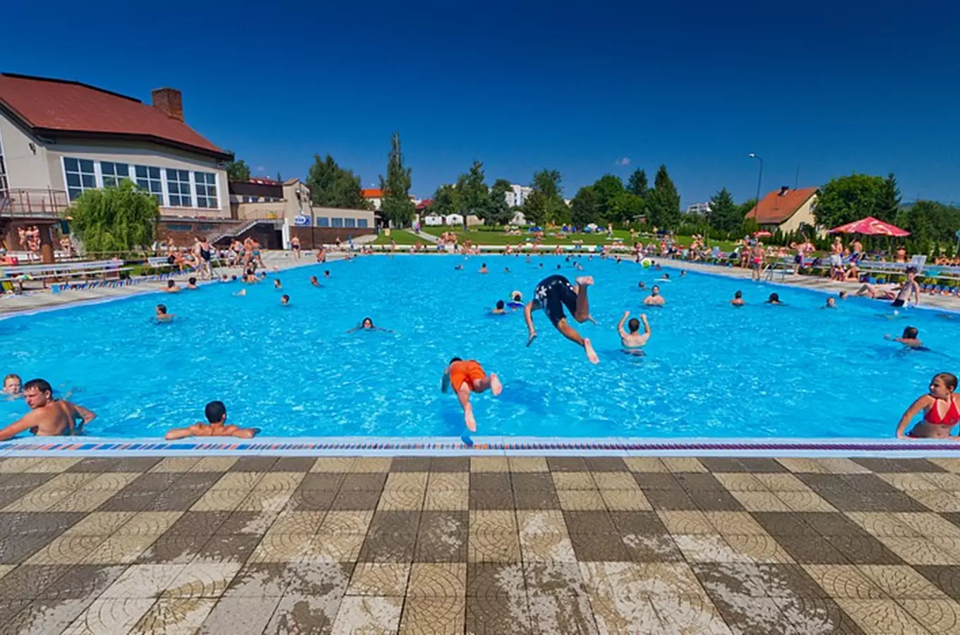 Lidé plavou v venkovním bazénu za slunečného dne.
