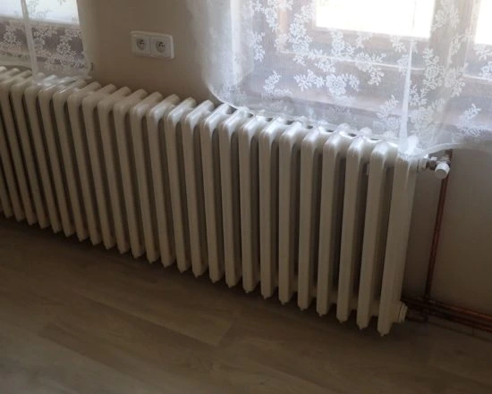Bílý litinový radiátor pod oknem s krajkovou záclonou.