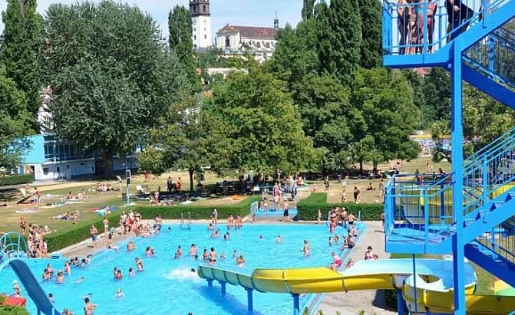 Lidé se koupejí v bazénu s vodními skluzavkami a historickým pozadím.