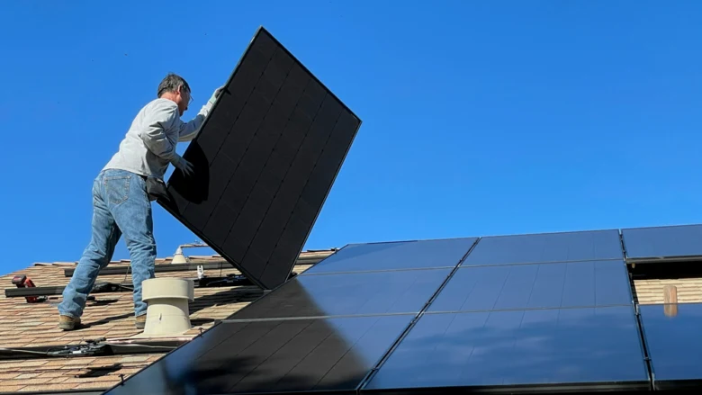 Muž montuje solární panel na střechu rodinného domu.