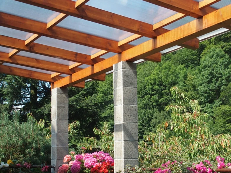 Pergola s průsvitnou polykarbonátovou střechou na pozadí zeleně.