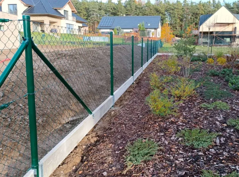 Zelená plot z pletiva kolem pozemku s domy v okolí.
