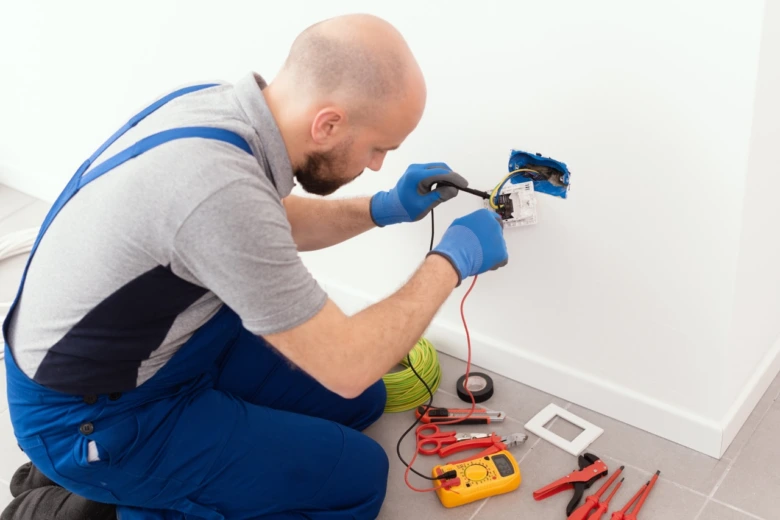 Elektrikář testuje instalaci v zásuvce multimetrem s nářadím na zemi.