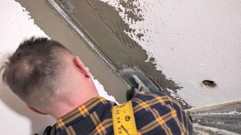 Muž lepi síťovou pásku na rohu stěny špachtlí.