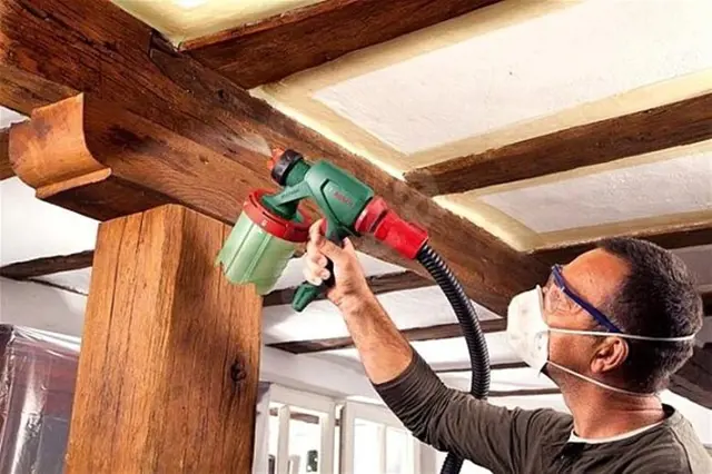 Muž stříká barvu na dřevěné trámy pomocí pistole.