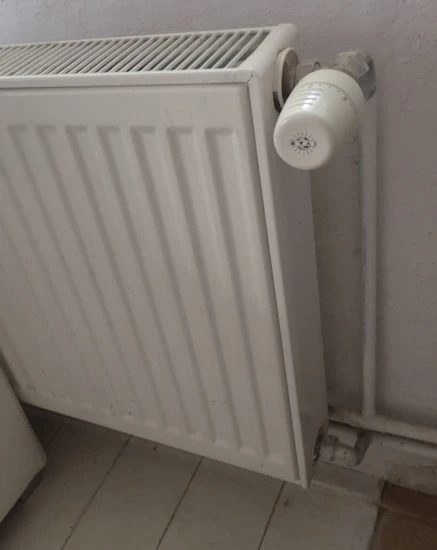 Bílý litinový radiátor připojený na potrubí u zdi.