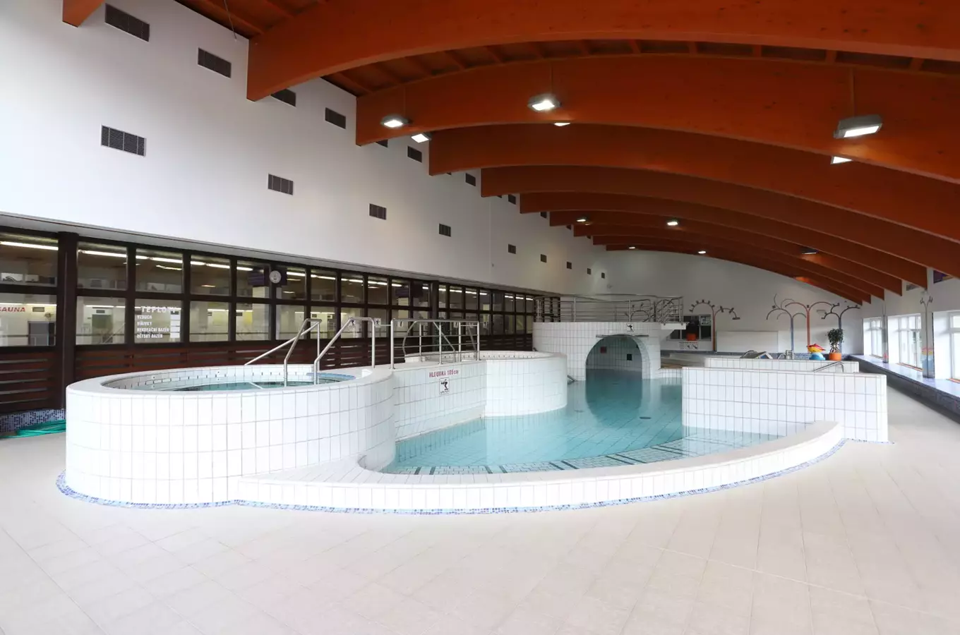 Vnitřní plavecký bazén s bílými dlaždicemi a dřevěným stropem.
