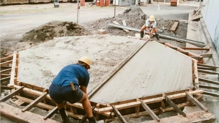 Dva dělníci nalévají beton do dřevěného patka pro dřevozstavbu.