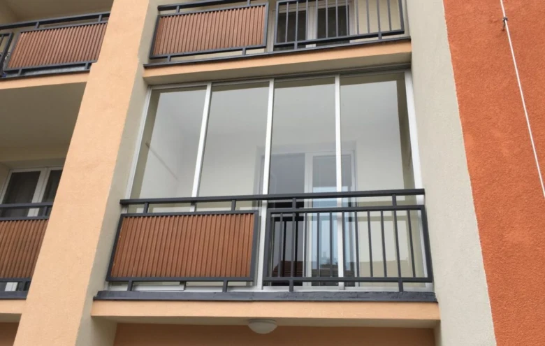 Nově zasklený balkon s černým zábradlím na oranžové fasádě.