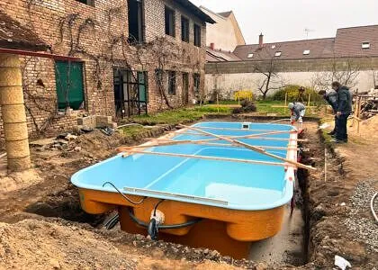 Modrý nadzemní bazén je instalován do vykopaného jámy vedle staré cihlové budovy.