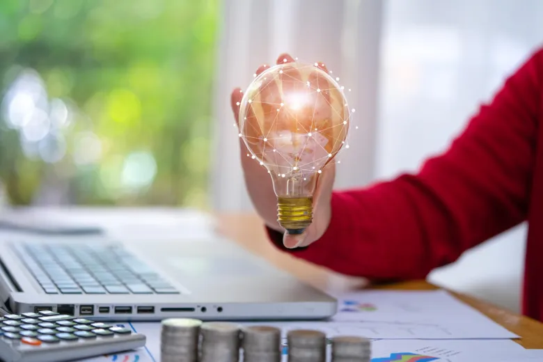 Ruka drží svítící žárovku s digitální mřížkou nad pracovním stolem.