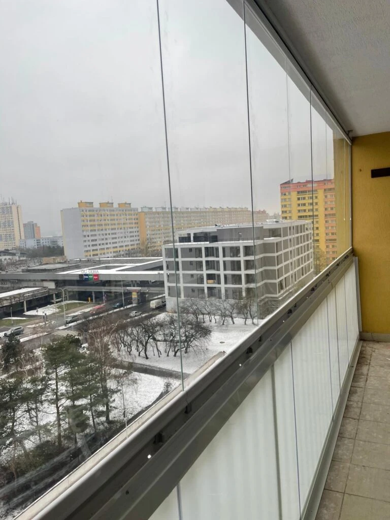 Zasklený balkon s výhledem na zasněženou ulici a budovy.