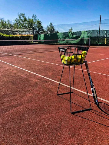 Koš na tenisové míče s raketou na červeném dvorci.