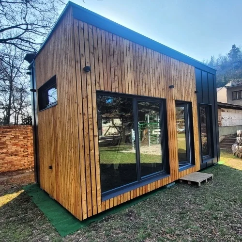Moderní dřevostavba malého domu s velkými okny na trávníku.